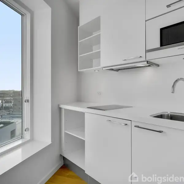 Et moderne køkken med hvidt skab og håndvask, i en lys lejlighed med en stor vinduesudsigt over bybygninger. Mikrobølgeovn indbygget i skabene.