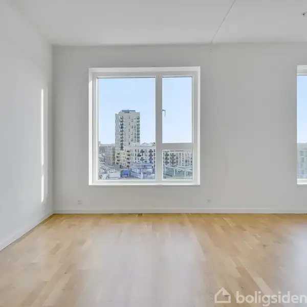 Et tomt værelse med lyse trægulve og hvide vægge har to store vinduer, der viser udsigt til moderne bygninger udenfor.