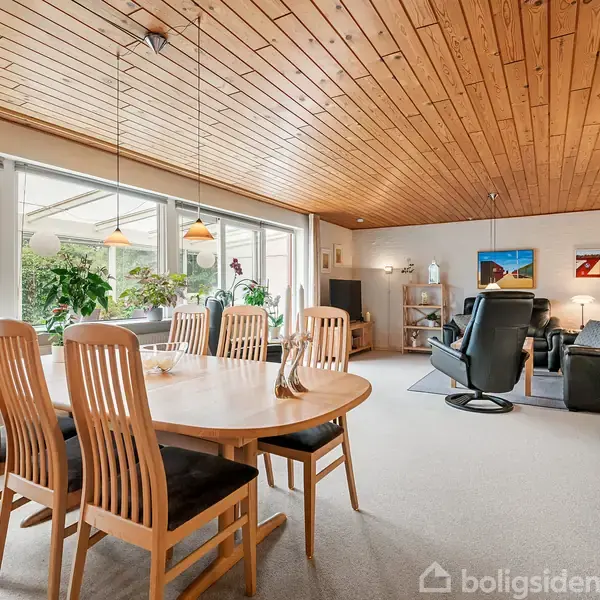 Spisebord med otte stole står i en stue med trægulv og træloft. Store vinduer slipper masser af lys ind og stuen har planter og billeder på væggene.
