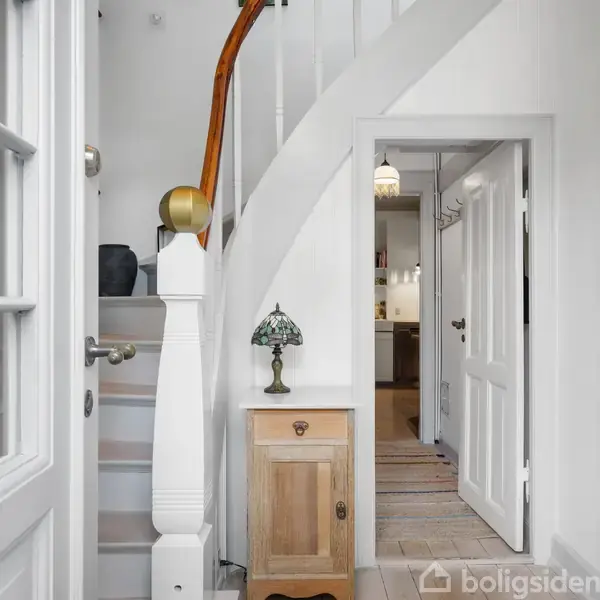 Trappeopgang med hvidmalede gelændere og trætrin; en lille kommode med en dekorativ lampe står ved siden af. Der ses en åbning mod et lyst andet rum med dør.