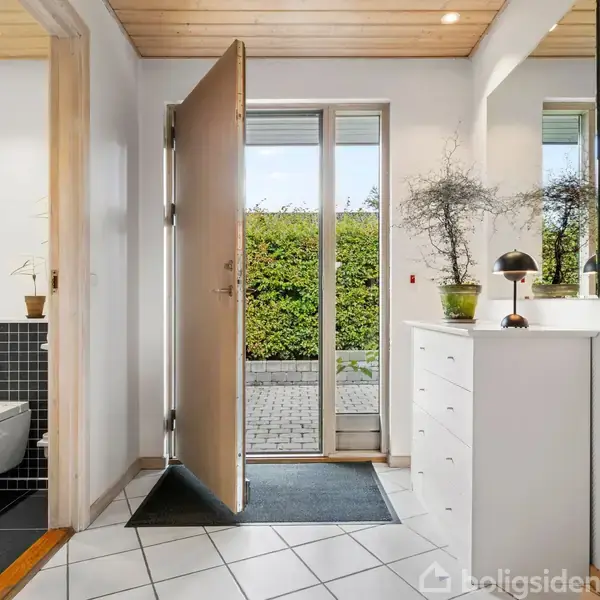 Dør står åben, fører til en flisebelagt indkørsel, omgivet af hække. Til venstre er et toilet med sorte fliser. Til højre en kommode med planter og en lampe.