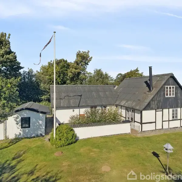 Et bindingsværkshus står i en have med græsplæne og træer. Et flag vajer på en stang ved siden af huset, omgivet af åbne marker og en klar blå himmel.