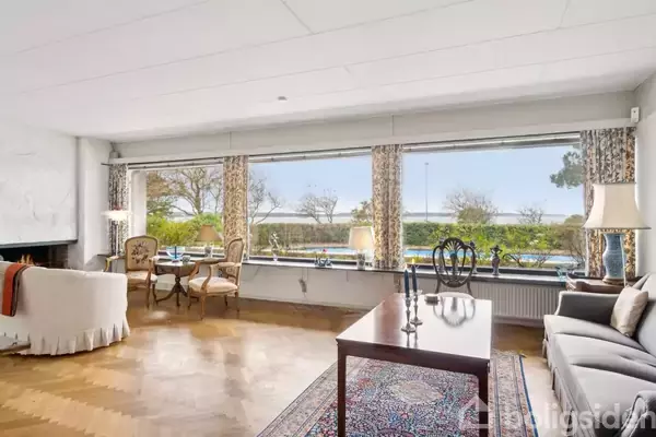 Stue med store vinduer tilbyder udsigt til havet. Møbler inkluderer en sofa, lænestole og et sofabord over et tæppe. Trægulv og pejs giver rummet en hyggelig atmosfære.