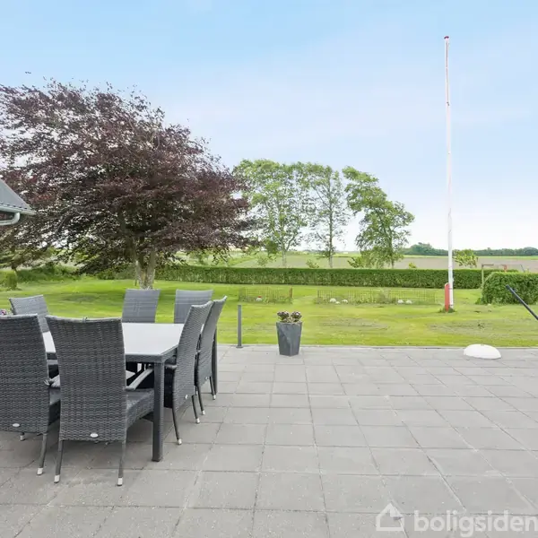 Udendørs terrasse med sort spisebord og seks stole placeret på flisebelægning. I baggrunden er der en græsplæne med træer og et flagstang i en landlig have.