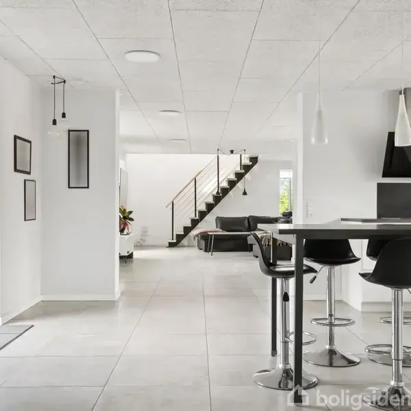 Et moderne køkken- og stueområde med sorte barstole ved en køkkenø. En trappe fører opad. Høje, hvide vægge med hængende lamper og kunst skaber et stilrent miljø.