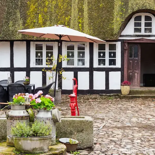 Stråtækt bindingsværkshus med små vinduer. Gårdsplads har et bord med parasol og stole. Blomster og en rød vandpumpe er midtpunkt, omgivet af stenbelægning. Ingen tekst til stede.