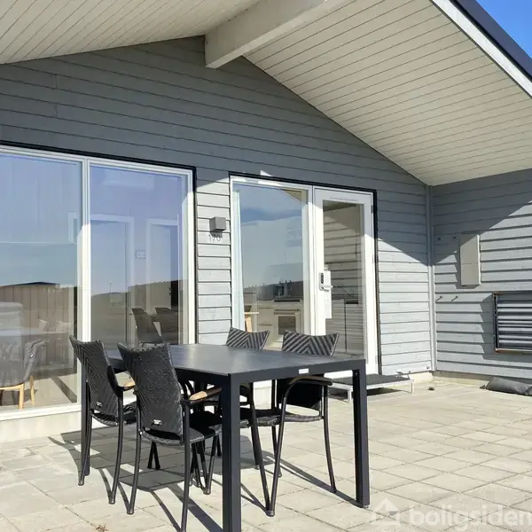 Terrassebord med fire stole står på en flisebelagt terrasse. I baggrunden er et moderne hus med store vinduer og en dør, omgivet af en solrig himmel og rolige omgivelser.