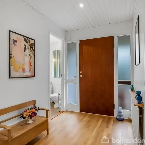 En entré med en trædør i midten, en bænk til venstre med blomster og et billede på væggen. Til højre ses en radiator og dekorationer.