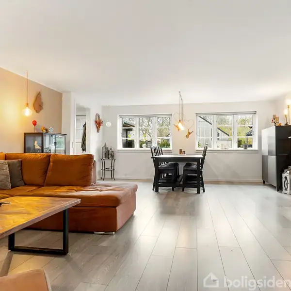 En brun hjørnesofa står i en lys stue med trægulv. Et træbord har dekorative lys. I baggrunden ses et spisebord med stole foran store vinduer.