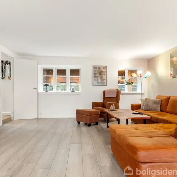 En stor orange sofa står i en stue ved et lavt træbord. TV står på en bænk til venstre. Lyse trægulve og hvide vægge med flere billeder.