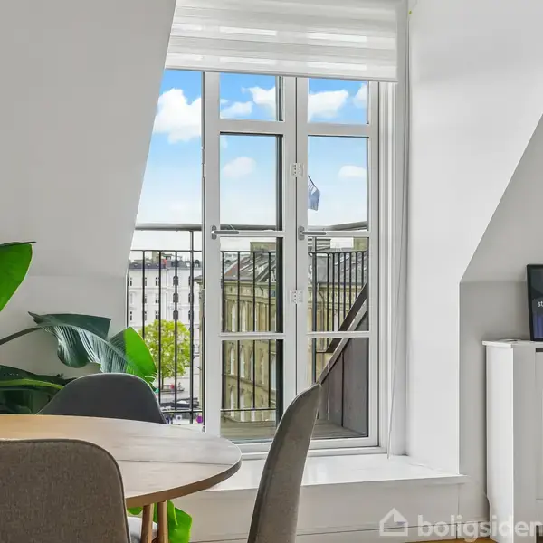 Et lyst rum med et rundt spisebord til venstre, omgivet af stole. Store grønne planter står ved siden af bordet. En dør fører ud til en balkon, og et skilt ved radiatoren siger "Facts are stubborn, statistics are more pliable."