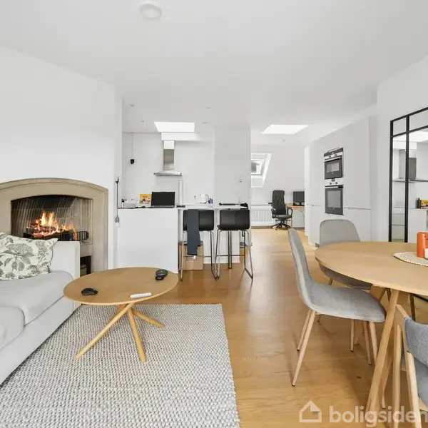 En stue med sofa og brændende pejs; sofabord og rundt spisebord med stole. I baggrunden ses et åbent køkken med barstole, og rummet er lyst og moderne indrettet.