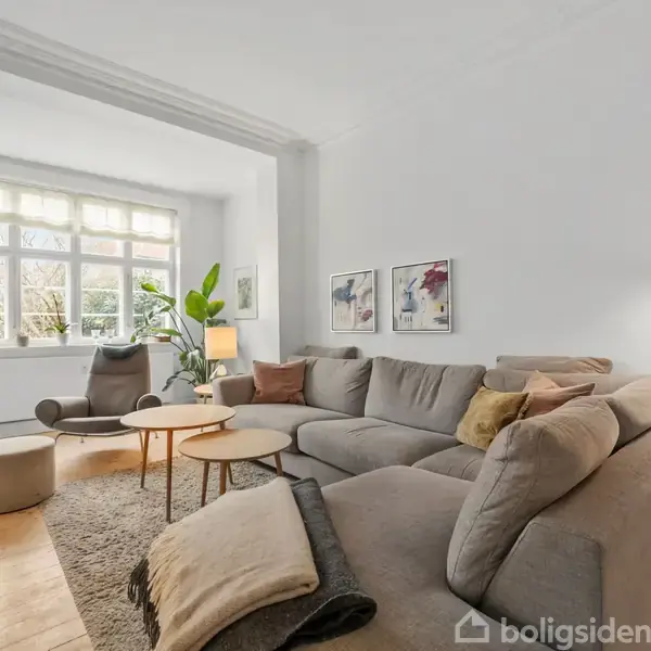 En beige hjørnesofa står i en lys stue ved siden af et sofabord og en grå lænestol. Rummet har hvide vægge, trægulv og store vinduer med planter.