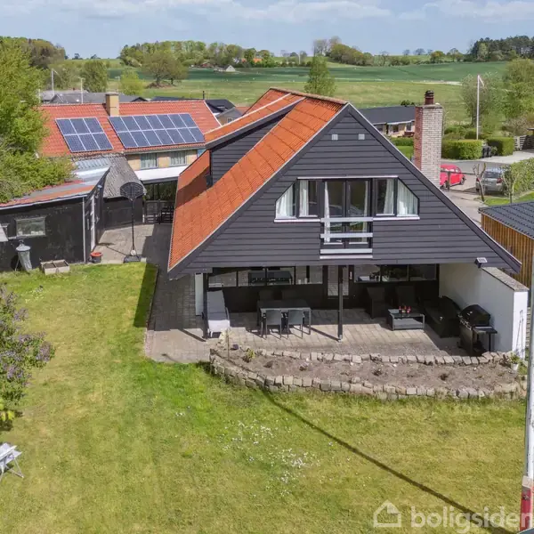 Hus med sort tag og stor terrasse; smukt omkranset af grøn græsplæne og træer, beliggende i et landligt område med naboer. Bænke ses i haven, solen skinner klart.