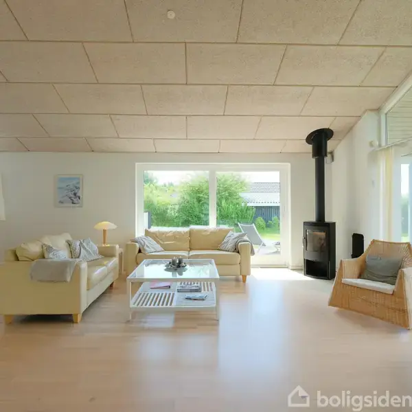 Stue med lys sofa og kurvestol omkring et sofabord. En brændeovn i hjørnet. Store vinduer giver udsigt til have. Spisebord med stole står til venstre.