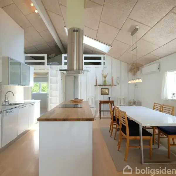 Køkken-alrum med moderne hvide skabe og apparater til venstre, et spisebord med træstole til højre, under et skrånende loft med åbne loftsbjælker og store vinduer i en lys indretning.