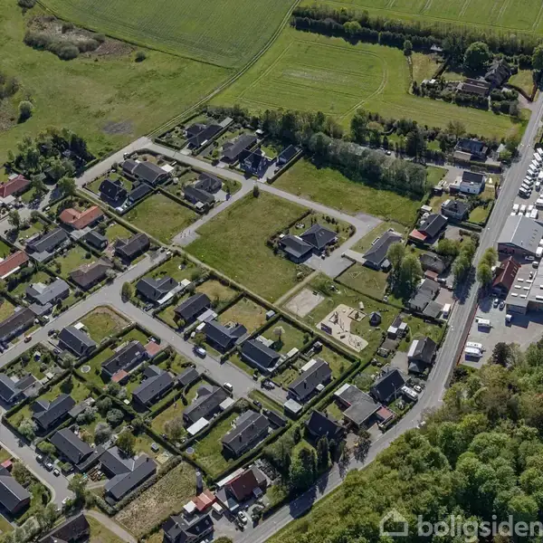 Et parcelhuskvarter set oppefra med rækker af huse, veje og indkørsler. Grønne områder og marker omkranser kvarteret. En vej med industribygninger og parkerede biler ligger til højre.