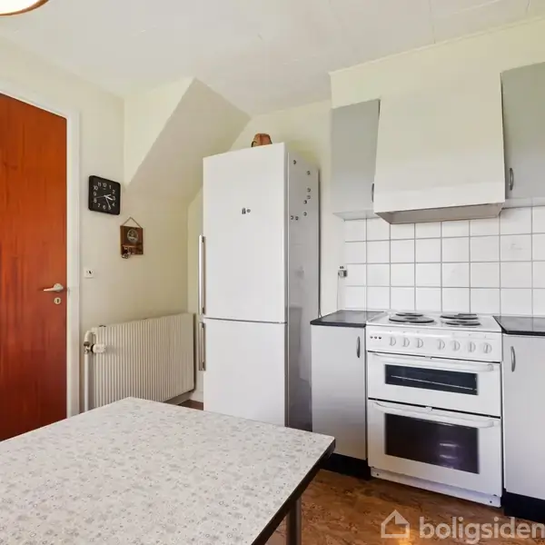 Et køkken med et køleskab ved siden af en ovn med kogeplader. Der er en køkkenbordplade i forgrunden, og en trædør samt vægur i baggrunden.