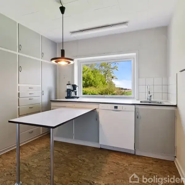 Et køkken med grå skabe og skuffer, en bordplade med kaffemaskine og en stor vask. Et vindue åbner ud til en grøn udsigt med træer udenfor.