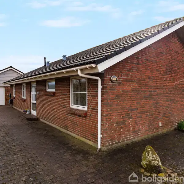 Rødt murstenshus med sort tag og hvide vinduer står på flisebelagt indkørsel, omgivet af en have. Garage ses i baggrunden under en blå himmel.