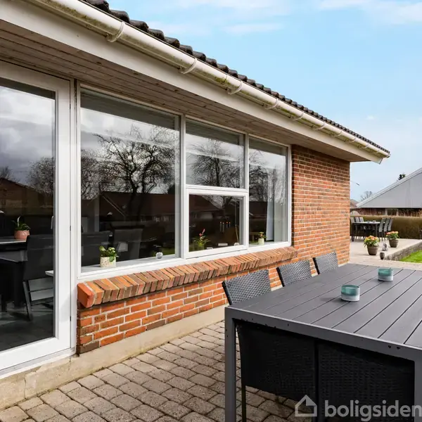Rundt bord med sorte stole står på en flisebelagt terrasse foran et murstenshus med store vinduer; haven ses i baggrunden med træer og et nabohus.
