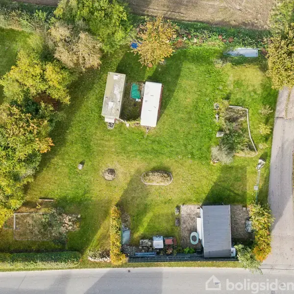 Grønt område med to små bygninger står stille, omgivet af træer og buske. En vej løber langs højre side, mens en sti fører til indgang i nederste kant.