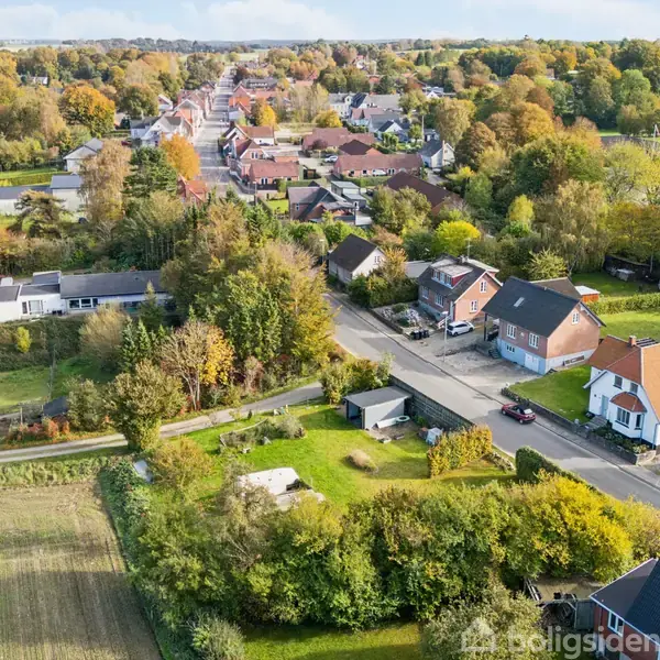 Byhus med rødt tag står ved en vej omgivet af grønne haver og træer. I baggrunden ses flere huse og en landsby i et frodigt, skovklædt område.