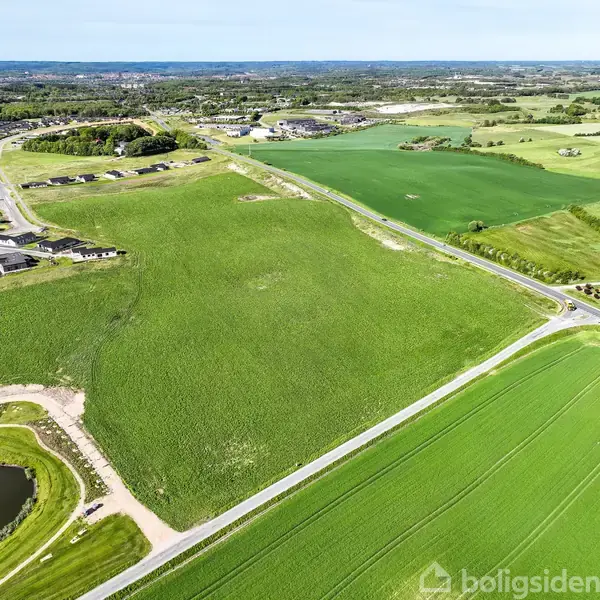 Grøn mark strækker sig langs veje, omgivet af boligområder og en dam til venstre. Bydelens bygninger ses i baggrunden. Området ser landligt og fredeligt ud.