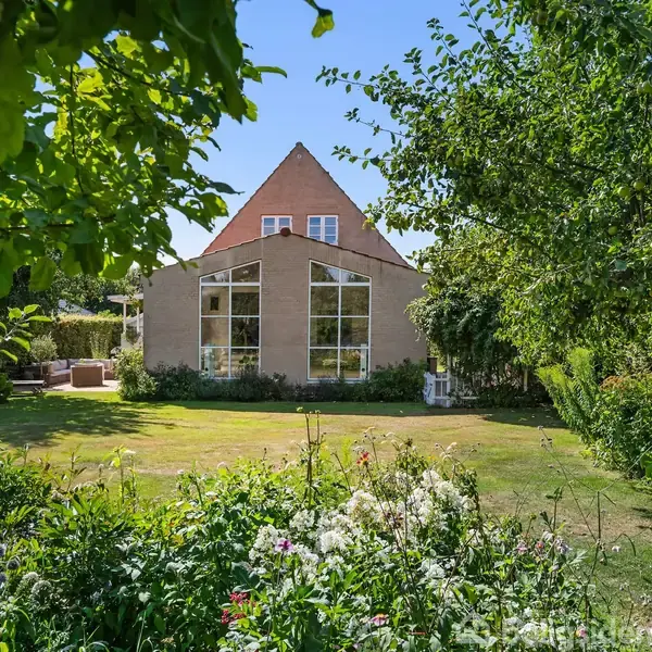 Et hus med skråt tag og store vinduer står stille omgivet af en frodig have med forskellige blomster og træer under en klar, blå himmel.