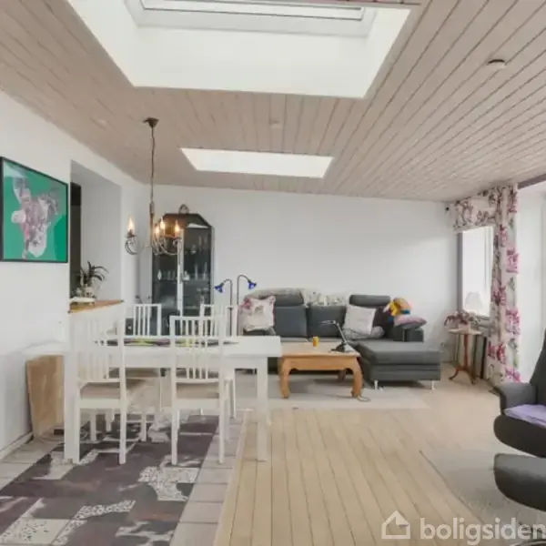 Stue med spisebord og sofagruppe. Spisebord står til venstre under tagvindue nær farverigt maleri, mens grå sofa og lænestol befinder sig til højre ved blomstrede gardiner.