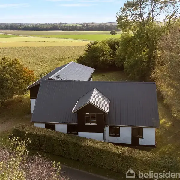En hvidmalet murstenshus med sort tag står stille i landskabet. Omgivet af grønne træer og marker i landlig idyl.