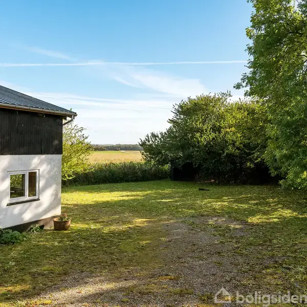Et hus står stille ved siden af en græsplæne, omringet af træer og marker under en blå himmel.