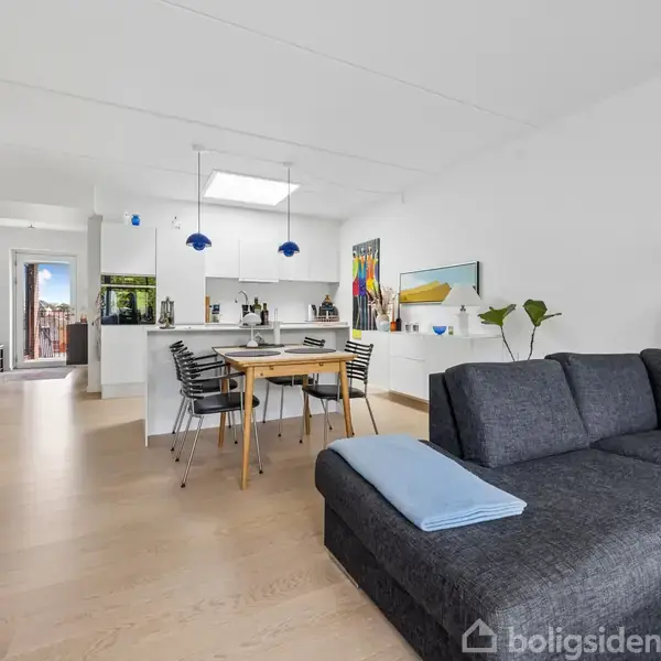 Grå sofa med blå pude står i en lys stue med trægulv. Bagved er et spisebord med stole og hængelamper. Køkkenområdet ses i baggrunden med moderne indretning.