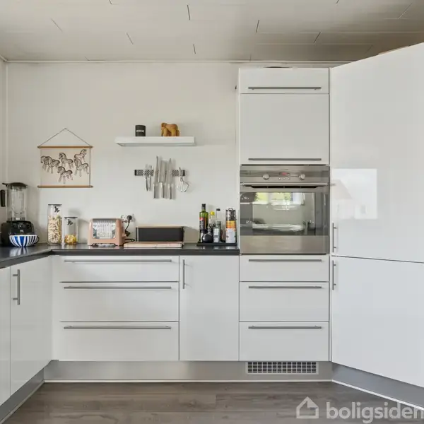 Et moderne køkken med hvide skabe og skuffer. På væggen hænger hylder med dekorationer. Et vindue lader lys strømme ind, og et stort køleskab står til højre.