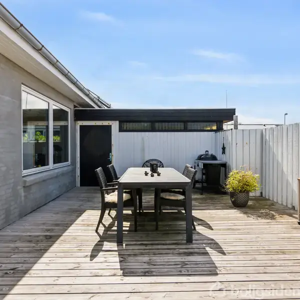 En træterrasse med et bord og fire stole står solbelyst. Omkring ses en lav mur og hegn, med blomsterkrukker og et lille bord til planter ved siden af. Ingen tekst er synlig.