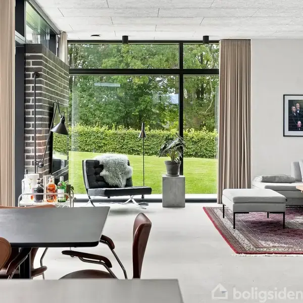 En moderne stue med sorte møbler og et tæppe. En stol ved et stort vindue med udsigt til en grøn have. En lampe og et bord med drikkevarer ved siden af.