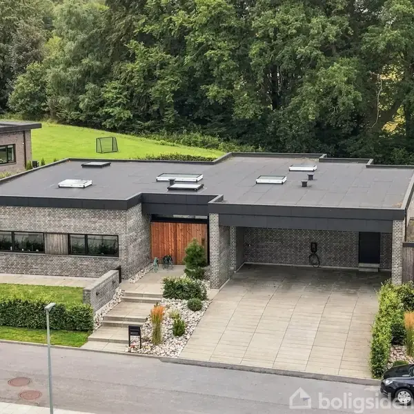 En moderne, fladtag-etplansvilla står ved en vej omgivet af grønne omgivelser. Foran huset er der en flisebelagt indkørsel og garage. Lækker have og nærliggende natur.