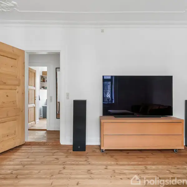 Fladskærms-TV står på en lav skænk, omgivet af højttalere i en stue med træparketgulv og åbning til anden del af lejligheden. Hvidmalede vægge og åbentstående dør.