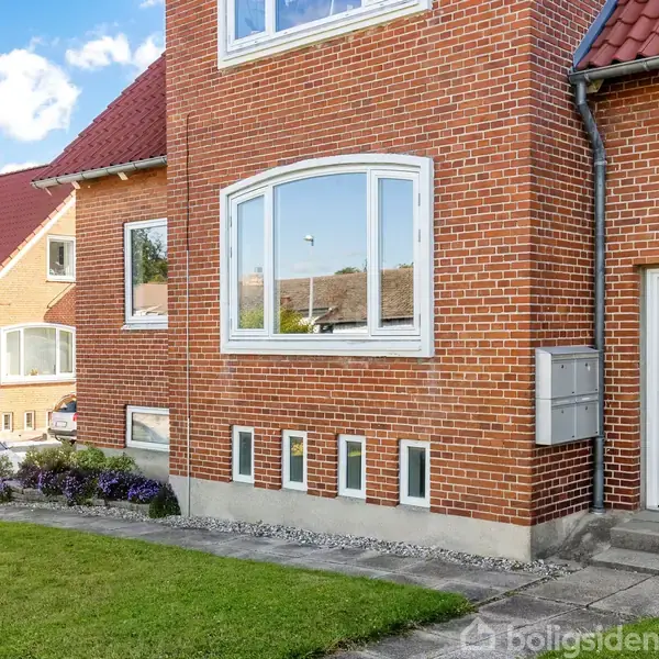 Rødt murstenshus med hvidmalede vinduer, nummer 16 ved indgangsdøren. Huset har grønne græsplæner og blomsterbede foran, og et nabohus kan ses i baggrunden.