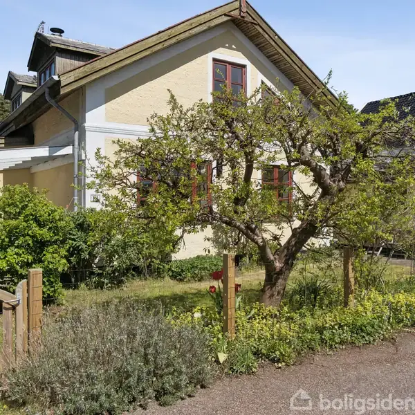 Hus i gul mursten med røde vinduer står bag et æbletræ i en lille have med grusvej foran og en træport i venstre side.