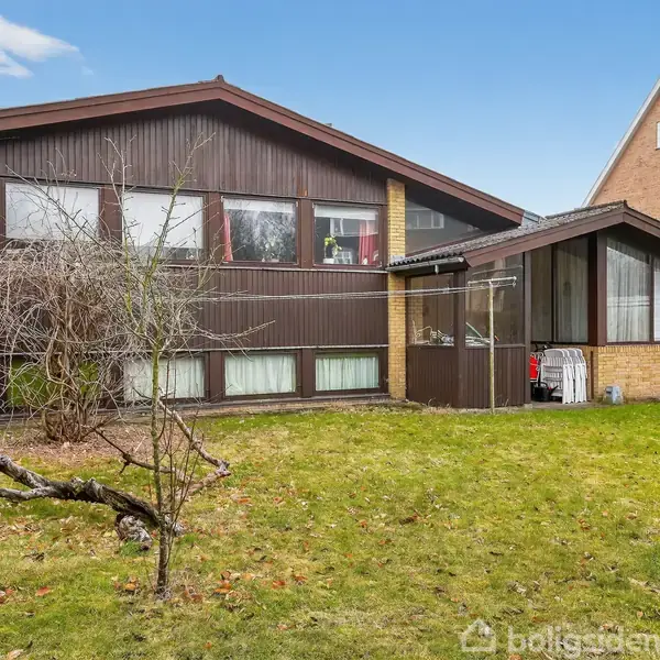 Et moderne træhus med store, mørke vinduespartier står i en grøn have, omgivet af hække. Ved siden af er et ældre hus med rødt tag. Himlen er blå med få skyer.