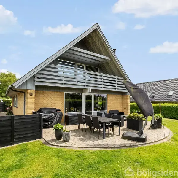 Et moderne hus har en spids tagkonstruktion. En terrasse med spisebord og parasol er placeret i en velplejet have. Himlen er delvist skyet.