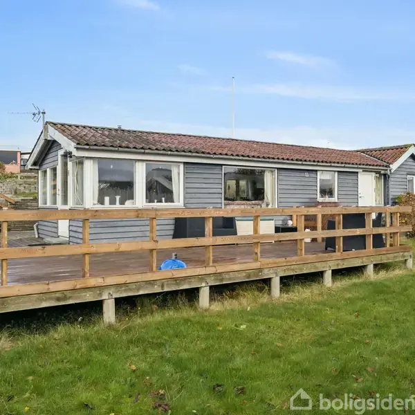 Et lille træhus med en træterrasse. Huset har store vinduer og er omgivet af en grøn græsplæne under en blå himmel.