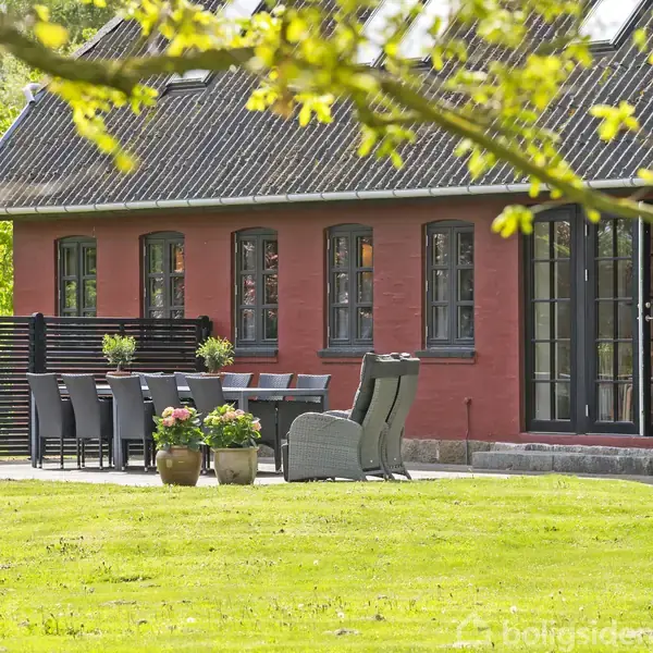 Rødt murstenshus med sorte vinduer. Havemøbler og blomsterkrukker står på terrasse. Omgivet af grøn have med træer og græs.