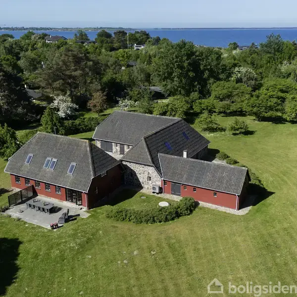 Et rødt hus med gråt tag står i en stor, grøn have med træer. Havet kan ses i baggrunden.