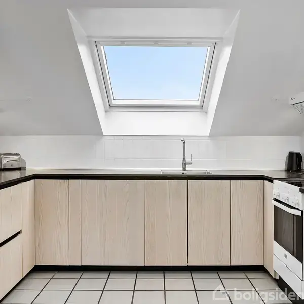 Køkken med lyse træskabe og sort bordplade. Køleskab og komfur står under et skråvindue, som lader naturligt lys ind. Hvidt flisegulv fuldender rummet.