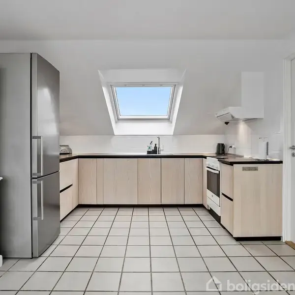 Et køkken med lyse træskabe og hvidevarer, herunder et køleskab og ovn, står i et rum med hvidt flisegulv. Et ovenlys sørger for naturligt lysindfald.