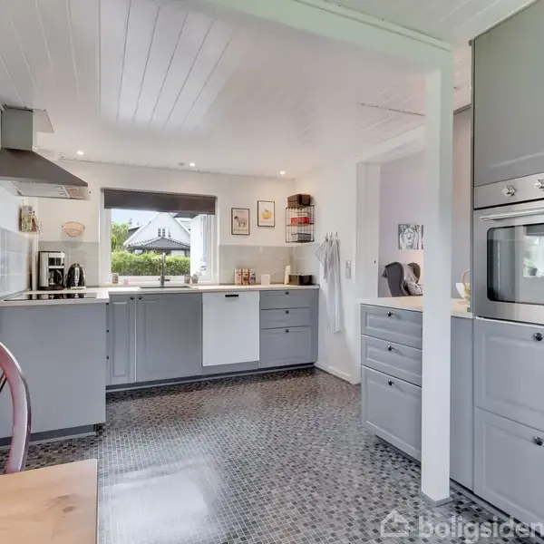 Køkken med grå skabe og integreret ovn, handling: stå stille, kontekst: moderne indretning med mosaikgulv, hvælvet loft og udsigt til have gennem vindue. Stol og bord i forgrund.