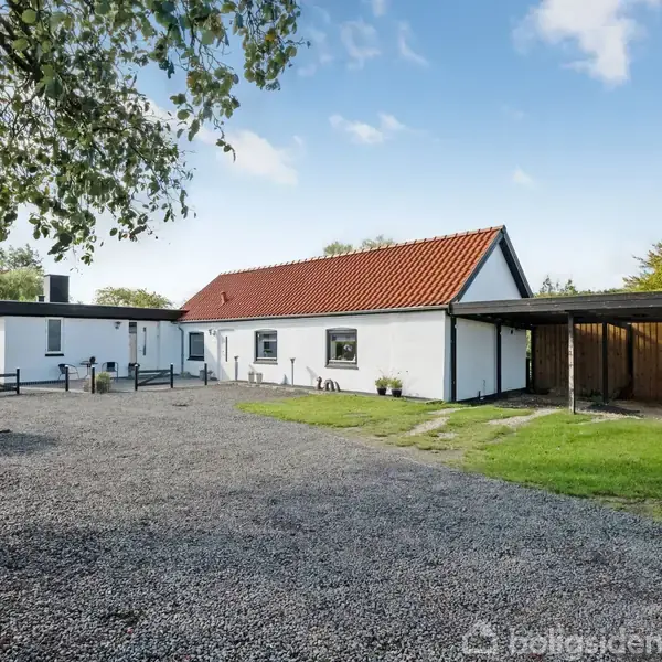 Hvidt hus med rødt tegltag står stille på en grusbelagt grund, omgivet af grøn beplantning og en åben carport til højre under en klar himmel.