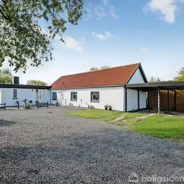 Et hvidt hus med rødt tegltag står stille ved siden af en carport. Gruset dækker indkørslen, omgivet af grønne buske og træer under en klar, blå himmel.